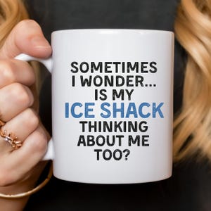 Pode incluir: Caneca de cerâmica branca com o texto "SOMETIMES I WONDER... IS MY ICE SHACK THINKING ABOUT ME TOO?" em preto e azul. A caneca tem uma alça curva e é segurada por uma pessoa.