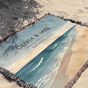 May include: A woven throw blanket featuring a beach scene with blue ocean waves and tan sand. The text "OLIVIA & JAKE" and "ENGAGED 2026" is printed in black on the upper left side. The blanket has a fringed edge with multi-colored threads.