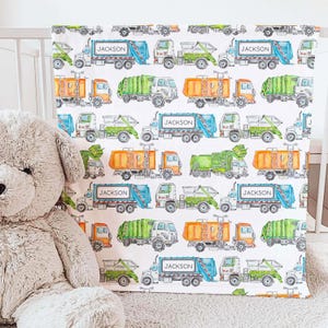 May include: A white baby blanket with a repeating pattern of blue, green, orange, and yellow garbage trucks. The trucks have the name "JACKSON" printed on the side.