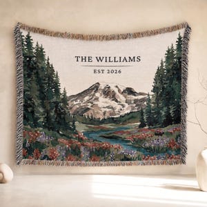 Puede incluir: Una manta tejida que muestra un paisaje de montaña con pinos, un río y flores silvestres. El texto THE WILLIAMS y EST 2026 está impreso en el centro. La manta tiene un borde con flecos y un patrón de hilos multicolores.