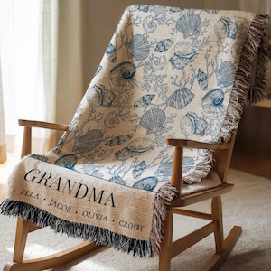 May include: A beige throw blanket featuring a blue seashell and coral design is draped over a wooden rocking chair. The blanket has a fringed edge and the word "GRANDMA" is printed in black, along with names.