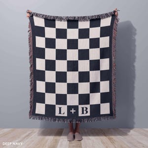 May include: Deep navy and cream-colored checkerboard throw blanket with fringe. The blanket features the initials "L + B" in white block letters at the bottom. The blanket is held up against a gray wall.