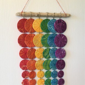 May include: A rainbow-colored wall hanging made of fabric circles strung on a driftwood branch. The circles are sewn with a radial pattern and are arranged in a rainbow order from red to purple.