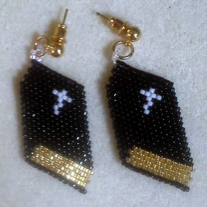 May include: A pair of black and gold earrings with a white cross design. The earrings are made of beads and have a diamond shape.