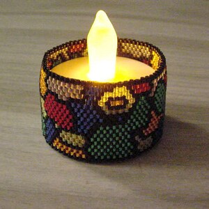 Puede incluir: Una vela decorativa con una envoltura de cuentas. La envoltura presenta un patrón colorido de cuentas rojas, amarillas, azules y verdes. La vela tiene un brillo suave y cálido, creando un ambiente acogedor.