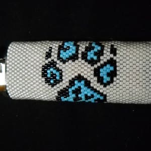May include: A white lighter cover with a blue and black paw print design. The cover is made of beaded material.