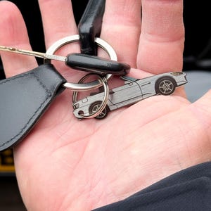 以下が含まれることがあります： コンバーチブルスポーツカーの形をした銀色の金属製キーホルダー。ホイール、ボディライン、ヘッドライトに黒い刻印が施されています。この車の形をしたチャームは、他の鍵と一緒に円形の金属製スプリットリングに取り付けられています。