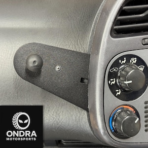 Puede incluir: Soporte de salpicadero de coche de metal negro con rótula para fijar accesorios. El soporte está fijado al salpicadero de un coche cerca de las rejillas de ventilación. El logotipo de ONDRA MOTORSPORTS está en la esquina inferior izquierda.