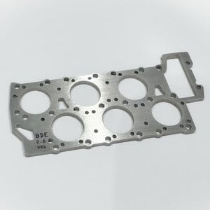 May include: A silver-colored metal component with multiple circular openings and smaller drilled holes. The component has a rectangular shape with a protruding section on one side. The text "BDE 2-8 VR6" is visible on the lower left corner.
