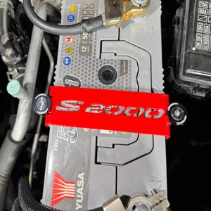 May include: A red metal battery tie-down bar with the text "S2000" cut out. The bar is secured with black fasteners. The battery is gray with the "YUASA" logo and a "12 Volt" label.