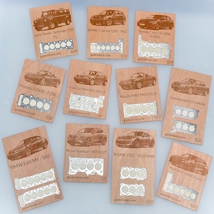 May include: A collection of eleven rectangular wooden plaques, each featuring a laser-engraved car model and a corresponding metal cylinder head gasket cutout. Text includes various vehicle names like Land Rover Defender, BMW, and Honda.