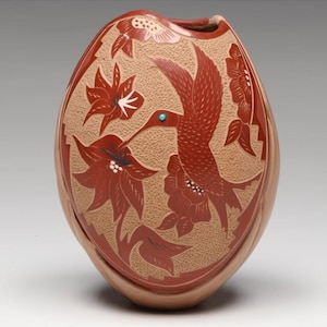 May include: A handcrafted, oval-shaped pottery vase with a textured tan background. The vase features a detailed design of a red hummingbird with a turquoise eye, surrounded by red flowers and leaves. The vase is a decorative piece.