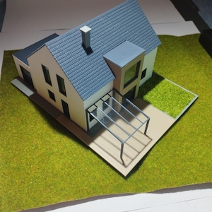 May include: A detailed architectural model of a modern house, featuring a grey tiled roof, white walls, and black window frames. The model includes a carport and is set on a green, grassy base, demonstrating contemporary design.