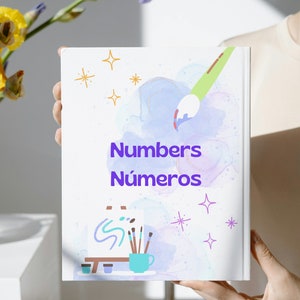 Puede incluir: Un libro blanco con un fondo de acuarela y una ilustración de pincel. El título del libro es "Numbers Números" en texto morado. La portada del libro también presenta un pequeño caballete con pinceles y una taza.