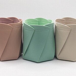 Könnte beinhalten: Drei dekorative Behälter aus Kunstleder. Die Behälter sind in den Farben Rosa, Grün und Beige gehalten. Jeder Behälter hat ein einzigartiges Falten-Design und eine genähte Naht am oberen Rand.
