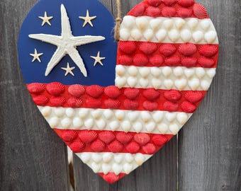 Op de afbeelding: Hartvormige patriottische wanddecoratie. Het ontwerp heeft een blauwe cirkel met een witte zeester en sterren, en een gedeelte met rood-witte schelpenstrepen. Er is een jute touw bevestigd om op te hangen.