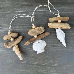 May include: Three driftwood ornaments with seashells and wooden beads. Each ornament is suspended from a white string. The ornaments are arranged in a row on a gray wooden surface.