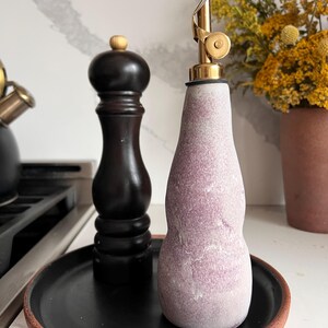 May include: A black pepper grinder and a purple and white oil dispenser with a gold spout sit on a black tray with a brown rim. Yellow flowers in a brown vase are in the background. The pepper grinder is dark brown with a wooden top.
