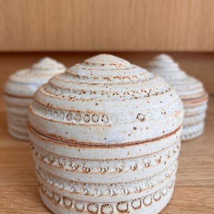May include: Three handmade ceramic lidded jars with a cream-colored glaze. The jars have a rounded shape with horizontal ridges and decorative patterns. The lids are conical, and the jars are arranged on a wooden surface.