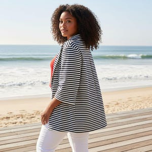 May include: A black and white striped blazer with three-quarter sleeves. The blazer is worn over a red top and white trousers. The background features a beach with the ocean and blue sky.