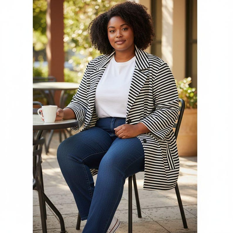 May include: A woman wearing a black and white striped blazer, white t-shirt, and dark blue jeans, seated at a table. She is holding a white mug. The blazer has a classic design with wide stripes. The setting appears to be outdoors.
