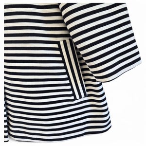May include: Close-up of a black and white striped jacket. The jacket features wide horizontal stripes and a pocket with vertical stripes. The sleeves are flared, and the fabric appears to have a textured surface.