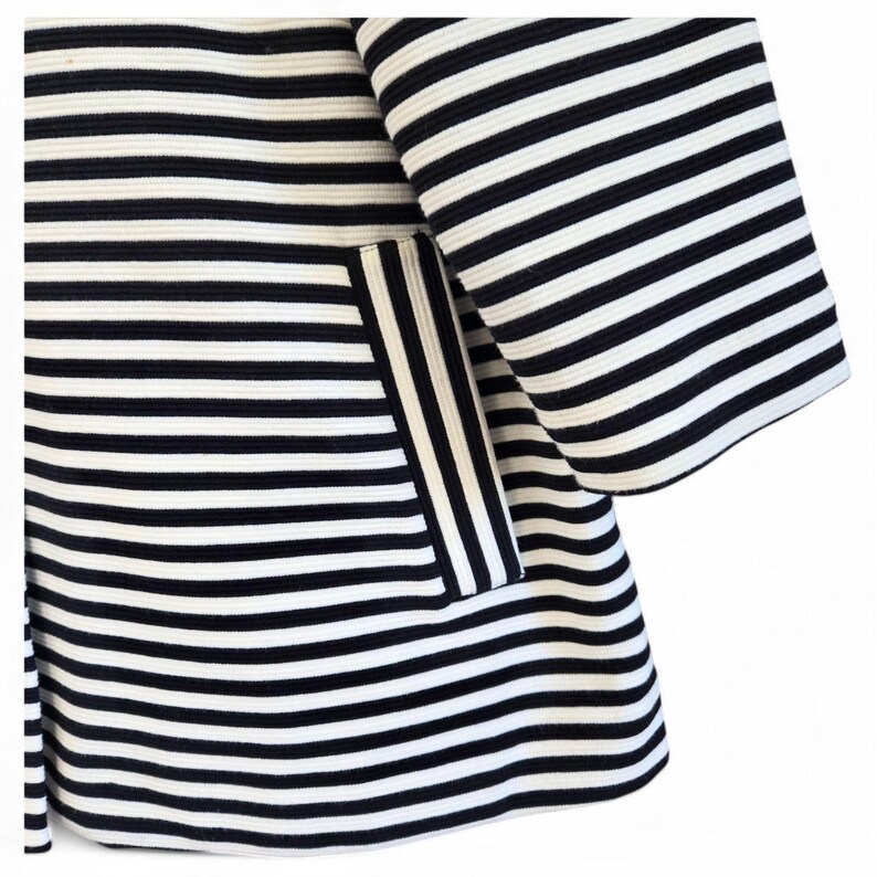 May include: Close-up of a black and white striped jacket. The jacket features wide horizontal stripes and a pocket with vertical stripes. The sleeves are flared, and the fabric appears to have a textured surface.