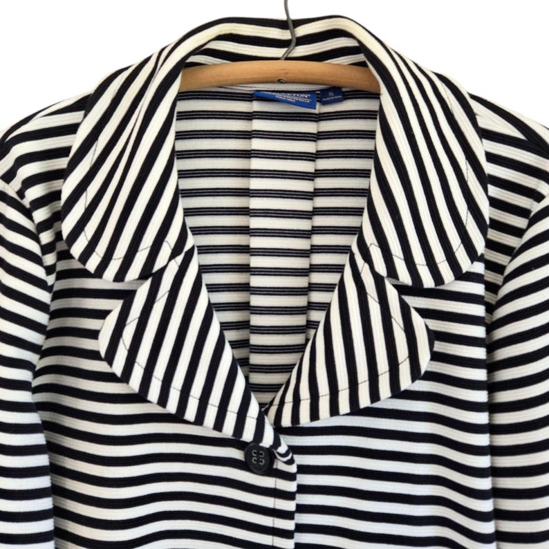 May include: A black and white striped blazer with a notched collar and a single button fastening. The jacket is suspended on a wooden hanger. The stripes are horizontal and evenly spaced.