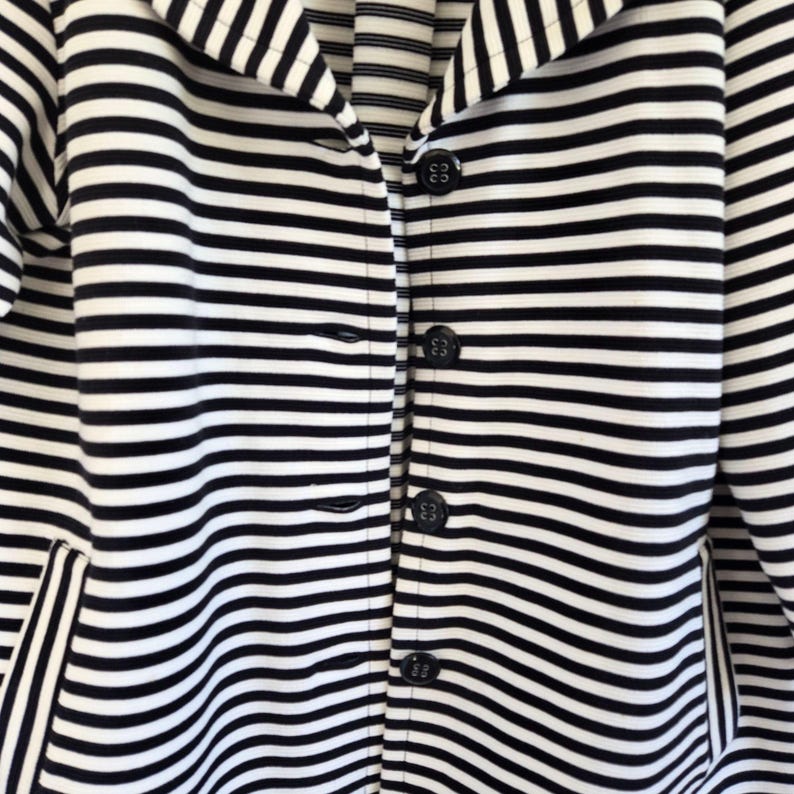 May include: A black and white striped blazer with a notched collar and four black buttons. The jacket has a classic design with horizontal stripes and a pocket on the left side. The fabric appears to be a medium weight.