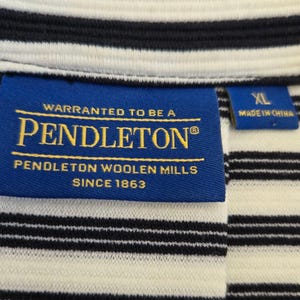 May include: Close-up of a striped shirt with a blue label that reads "PENDLETON WOOLEN MILLS SINCE 1863". The shirt has black and white horizontal stripes. The label also says "WARRANTED TO BE A PENDLETON".