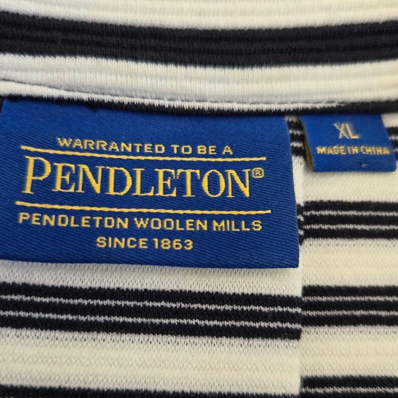 May include: Close-up of a striped shirt with a blue label that reads "PENDLETON WOOLEN MILLS SINCE 1863". The shirt has black and white horizontal stripes. The label also says "WARRANTED TO BE A PENDLETON".
