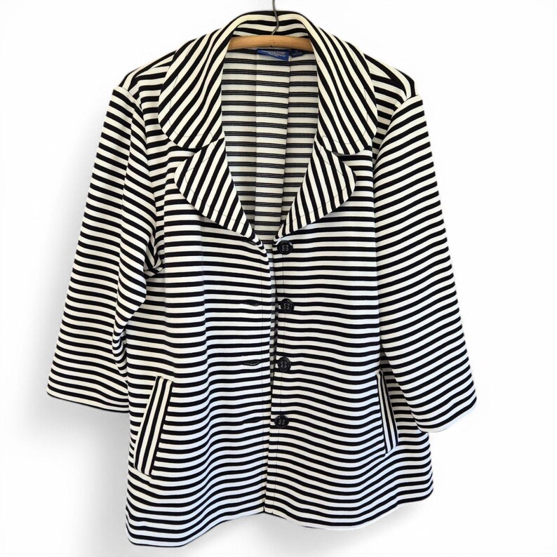 May include: A black and white striped blazer with three-quarter sleeves. The blazer features a notched collar, a three-button closure, and two front pockets. The garment is suspended on a wooden hanger.