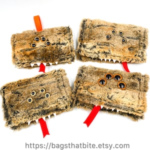 May include: Four brown and black faux fur monster pouches with black button eyes, white teeth, and red ribbon tongues. One pouch has an orange ribbon. The pouches are arranged on a white background.