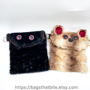 Puede incluir: Dos bolsas de felpa y peludas. Una es negra con detalles en rosa y ojos grandes con bordes rosas. La otra es de color canela con detalles en borgoña, ojos grandes y una nariz, que se asemeja a un oso. Ambas tienen anillas en D metálicas y el texto "https://bagsthatbite.etsy.com".