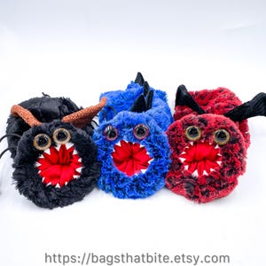 May include: Three plush monster pouches in black, blue, and red. Each has large googly eyes, a red mouth with white teeth, and various horn-like appendages. The pouches are fluffy and have a cartoonish design.