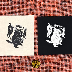 May include: Two square fabric patches featuring a badger illustration. One patch is off-white with a black badger holding a flag with a star, and the other is black with a white badger holding the same flag. The text Stabler Cake is at the bottom.