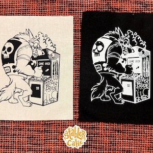 May include: Two square patches, one black and one beige, each featuring a white illustration of a wolf-like character playing an arcade game. The game machine has the words "GAME OVER" and "BEAST MODE". A sticker with the text "Stabler Cake" is also visible.
