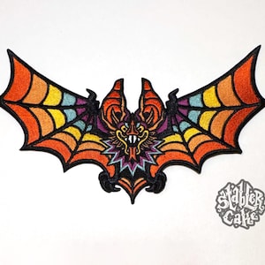 May include: Embroidered bat patch with a colorful design. The bat's wings feature a gradient of orange, yellow, and blue, with a black outline. The bat's face is detailed with orange, purple, and yellow. The patch has a black border and is on a white background.