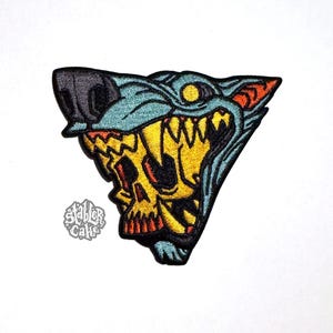 May include: Embroidered patch featuring a wolf's head with a skull design. The wolf is depicted in shades of blue, yellow, and orange, with sharp teeth and a menacing expression. The patch has a black border and the text "Stabler Cake".