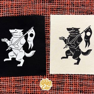 May include: Two square patches with a wolf design. One patch is black with a white wolf holding a flag, the other is off-white with a black wolf. The patches are on a red and orange patterned fabric.