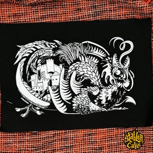 May include: A rectangular black fabric patch featuring a white screen-printed illustration of a mythical creature with wings and a long tail coiled around a castle. The text STABLER CAKE is printed in the bottom right corner of the patch.