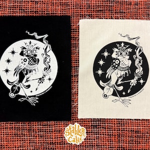 May include: Two square patches featuring a black and white owl design. The owl is inside a circle with stars, holding a torch. One patch is black, the other is off-white. The patches are on a red and orange patterned fabric.