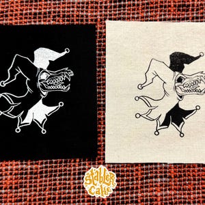 May include: Two square patches featuring a black and white jester design with a skull-like face. One patch is black, the other is off-white. The patches are on a patterned orange and brown surface. The patches are likely for clothing.