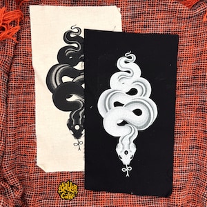 May include: Two rectangular fabric patches featuring a coiled snake illustration. One patch is off-white with a black snake, and the other is black with a white snake. Both snakes have a third eye on their heads and a knotted tongue. A small logo reads Stabler Cake.