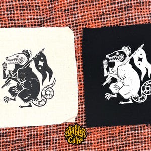 May include: Two square fabric patches featuring a screen-printed opossum holding a flag with a star symbol. One patch is off-white with a black print, and the other is black with a white print. The text Stabler Cake is printed in yellow at the bottom.
