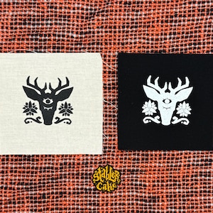 May include: Two square fabric patches featuring a stylized deer head with a central third eye and floral accents. One patch is off-white with a black print, and the other is black with a white print. A small logo with the text Stabler Cake is below.