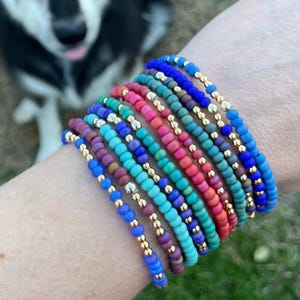 May include: A stack of ten beaded bracelets worn on a wrist, featuring small round beads in various colors including blue, turquoise, pink, and purple, interspersed with small metallic gold-toned spacer beads throughout each individual strand.