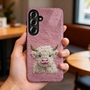 May include: A pink phone case with a textured surface and a detailed embroidered image of a Highland cow. The cow has white fur, a pink nose, and small green horns. The phone case has a triple-lens camera.