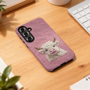 May include: A dusty rose phone case featuring a textured surface and a detailed embroidered image of a Highland cow. The cow has white fur, green horns, and a pink nose and mouth. The phone case has a black border.