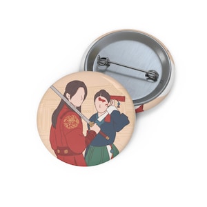 Könnte beinhalten: Ein runder Ansteckbutton mit einer digitalen Illustration von zwei Figuren in traditioneller Kleidung. Eine Figur trägt ein rotes Gewand mit Goldstickerei und hält ein Schwert, während die andere ein blau-grünes Outfit mit rotem Haaraccessoire trägt.